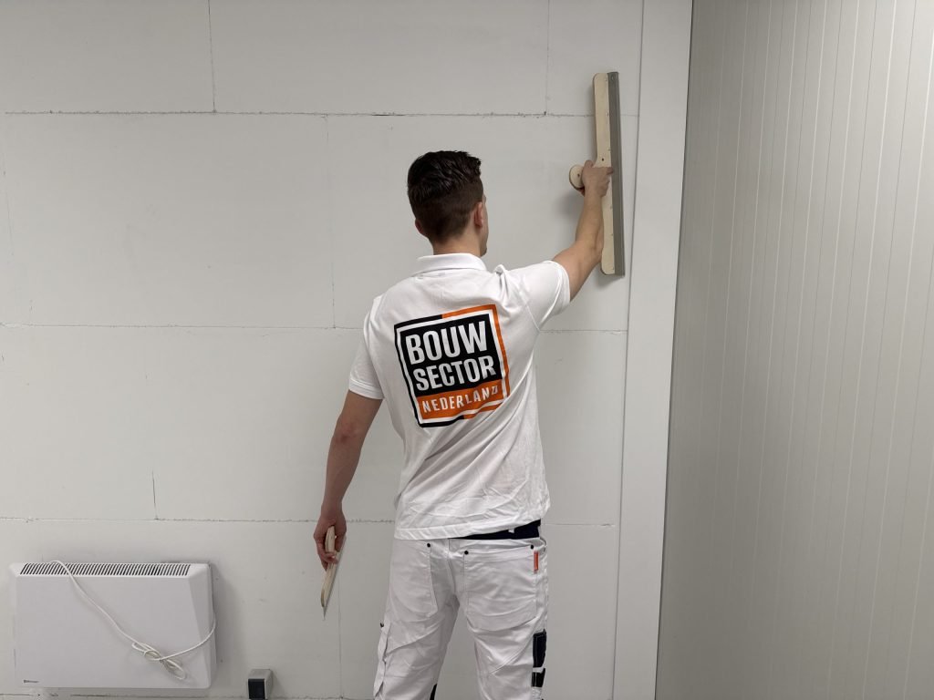 Stukadoor met het Bouwsector-logo op zijn rug zet een brede spaan boven zijn schouder tegen een witte wand