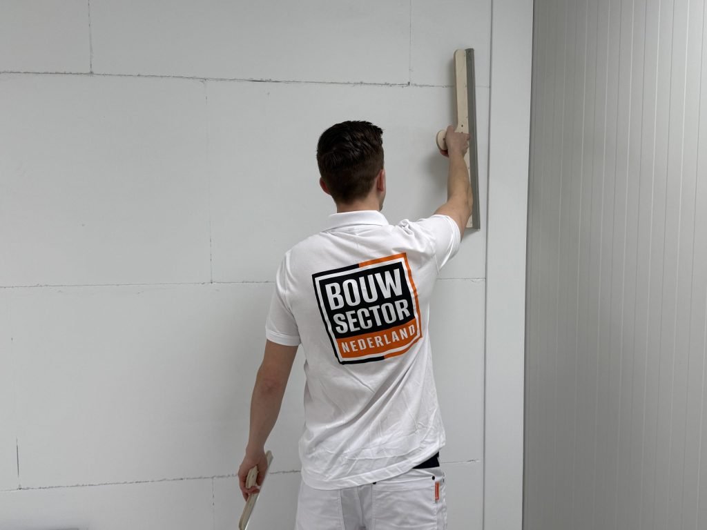 Stukadoor houdt een lange spaan rechtop tegen een wand van grote witte platen