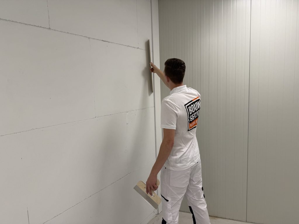Stukadoor trekt met een lange spaan een wand vlak in een lege gang met een roldeur ernaast