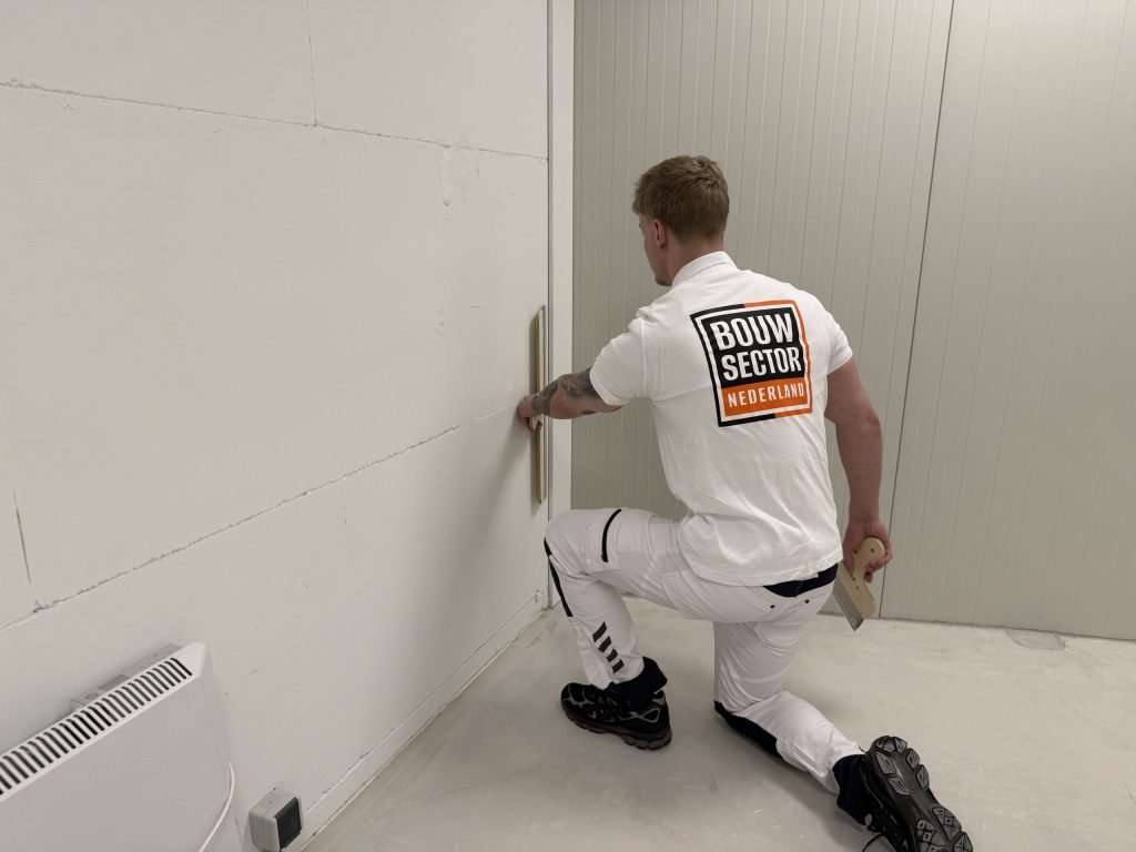 Stukadoor zit op één knie en trekt met een spaan het onderste deel van een wand naast de deurpost vlak