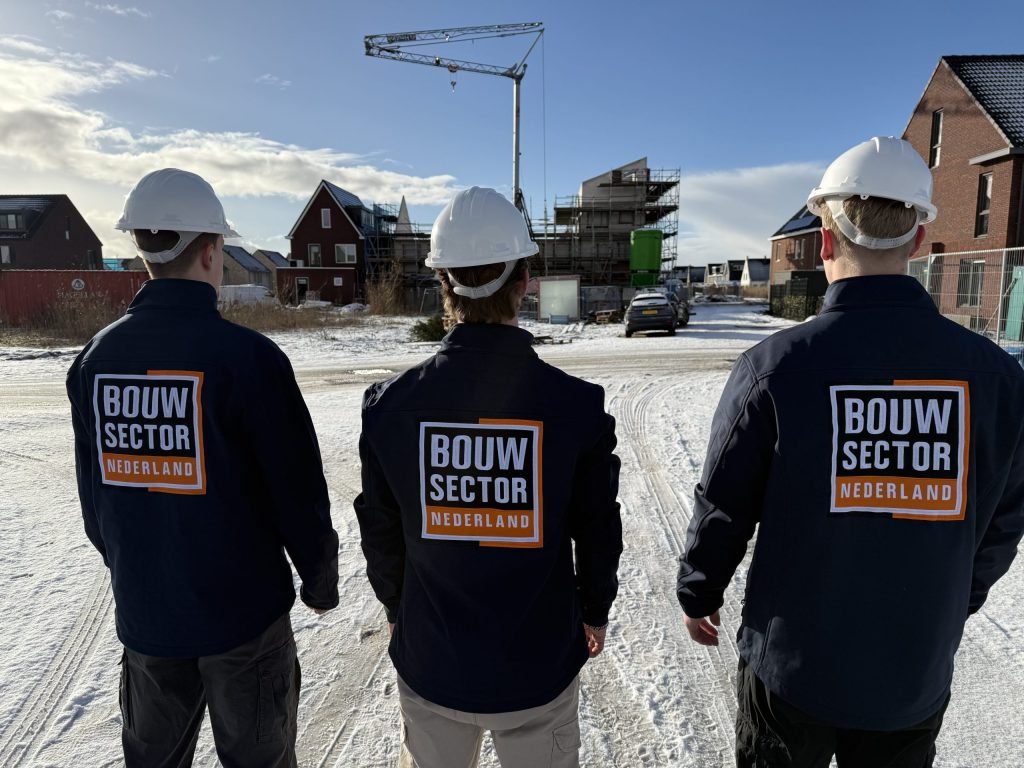 Drie vakmensen in werkjassen van Bouwsector Nederland met witte helm kijken naar een straat met woningen in aanbouw
